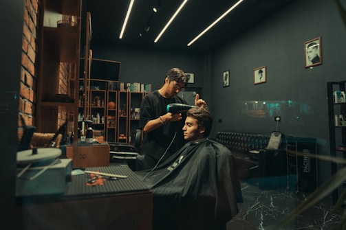 A modern barbershop with a stylish interior featuring exposed brick walls, sleek shelving filled with products, and framed photos on the walls. A barber is attentively working on a customer's haircut, using a hairdryer. The atmosphere is cozy and well-organized.