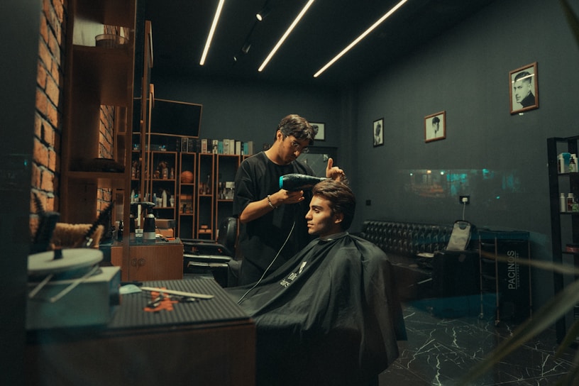 A modern barbershop with a stylish interior featuring exposed brick walls, sleek shelving filled with products, and framed photos on the walls. A barber is attentively working on a customer's haircut, using a hairdryer. The atmosphere is cozy and well-organized.