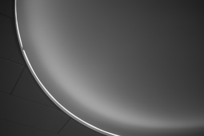A minimalist design featuring a smooth, curved surface with a gradient from dark to light gray, accented by a thin, bright white line that separates the gradient. The background consists of dark geometric shapes, possibly ceiling tiles.