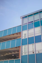 A multi-story building with a modern facade featuring large blue panels and metal frames. The structure has a combination of open parking levels and windows, with visible concrete beams and a brick column. The building is set against a clear blue sky.