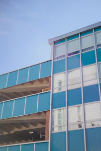 A multi-story building with a modern facade featuring large blue panels and metal frames. The structure has a combination of open parking levels and windows, with visible concrete beams and a brick column. The building is set against a clear blue sky.