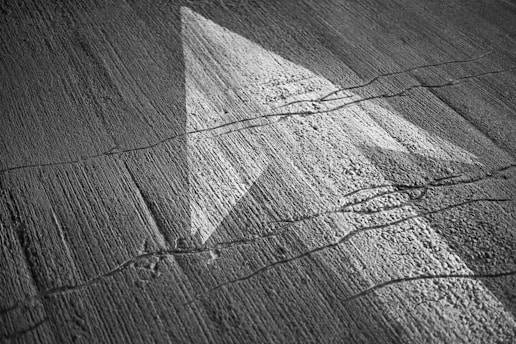 A textured concrete surface with a white arrow pointing forward. Cracks and lines create an intricate pattern on the ground.