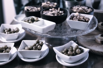 A selection of black and green olives is displayed in several small white square bowls on a glass tray. In the background, there are additional black bowls filled with an assortment of mixed nuts, including hazelnuts and almonds. The setting appears to be an elegant dining presentation.