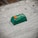 A small green electronic component or device placed on a rough surface. The component has orange and white markings, with text that reads 'PB 08G300 CA07 01006'. The surface appears to be made of concrete or a similar material and has some dark spots and imperfections.