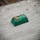 A small green electronic component or device placed on a rough surface. The component has orange and white markings, with text that reads 'PB 08G300 CA07 01006'. The surface appears to be made of concrete or a similar material and has some dark spots and imperfections.