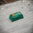 A small green electronic component or device placed on a rough surface. The component has orange and white markings, with text that reads 'PB 08G300 CA07 01006'. The surface appears to be made of concrete or a similar material and has some dark spots and imperfections.