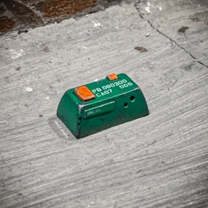 A small green electronic component or device placed on a rough surface. The component has orange and white markings, with text that reads 'PB 08G300 CA07 01006'. The surface appears to be made of concrete or a similar material and has some dark spots and imperfections.