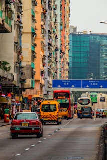 A busy urban street lined with tall, colorful apartment buildings on the left side. Various vehicles, including buses, vans, and cars, are seen on the road. The background features a large, modern glass building, and a blue road sign indicates directions to Mong Kok and Kowloon City.