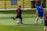 Two soccer players are on a grass field, one wearing a black uniform with the number 10 and the other in a blue uniform with the number 5. The player in black is kicking a soccer ball, while the player in blue is approaching him from behind. The field is surrounded by a fence, and there are trash bins in the background.