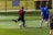 Two soccer players are on a grass field, one wearing a black uniform with the number 10 and the other in a blue uniform with the number 5. The player in black is kicking a soccer ball, while the player in blue is approaching him from behind. The field is surrounded by a fence, and there are trash bins in the background.