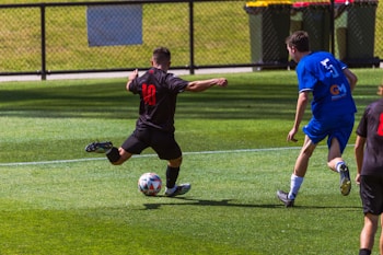 Two soccer players are on a grass field, one wearing a black uniform with the number 10 and the other in a blue uniform with the number 5. The player in black is kicking a soccer ball, while the player in blue is approaching him from behind. The field is surrounded by a fence, and there are trash bins in the background.