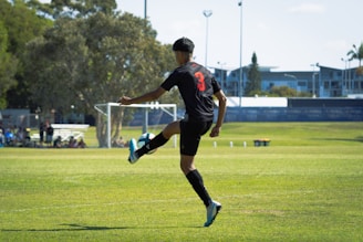 A soccer player is captured in mid-air action on a grassy field, wearing a black jersey with the number 3 visible, and black shorts. The player is in motion, appearing to kick the ball, which is slightly blurred to suggest movement. In the background, there are visible goalposts, trees, and a few buildings, with bright daylight illuminating the scene.