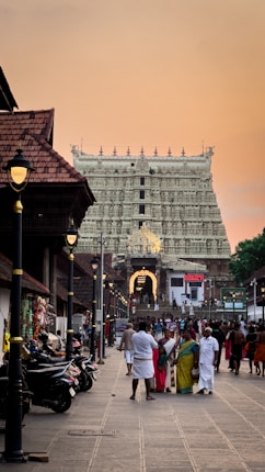 A bustling street scene in front of an intricately carved temple with a richly detailed facade, illuminated by soft lighting. The foreground features people walking along a pathway bordered by motorbikes, with traditional buildings lining the sides. The sky is a warm orange, suggesting either dawn or dusk.