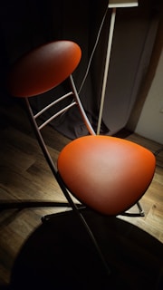 A modern chair with a sleek design featuring an orange-cushioned seat and backrest. The chair is placed on a wooden floor with soft lighting casting shadows around it. A standing lamp near the chair provides a warm glow, creating a cozy atmosphere.
