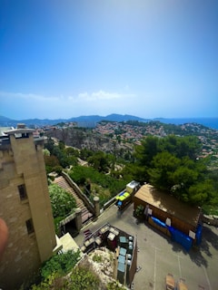 A panoramic view featuring a hilly landscape and a town with clustered houses. In the foreground, there is a section of an old stone building with stairs leading down and bright green foliage surrounding it. Further in the background, the town expands over the hills, and the sea is visible on the horizon under a clear, bright sky.