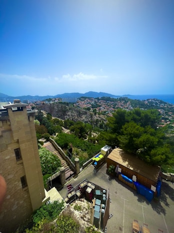 A panoramic view featuring a hilly landscape and a town with clustered houses. In the foreground, there is a section of an old stone building with stairs leading down and bright green foliage surrounding it. Further in the background, the town expands over the hills, and the sea is visible on the horizon under a clear, bright sky.