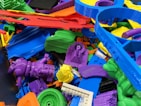 A collection of colorful 3D printed plastic objects in various shapes and colors, piled together. The objects include intricate designs such as gears, abstract forms, and tools, made from materials resembling different parts and components.