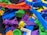 A collection of colorful 3D printed plastic objects in various shapes and colors, piled together. The objects include intricate designs such as gears, abstract forms, and tools, made from materials resembling different parts and components.