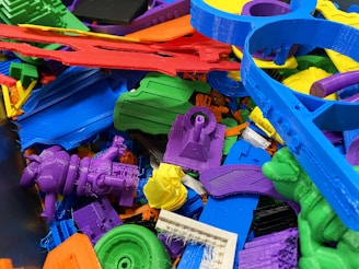 A collection of colorful 3D printed plastic objects in various shapes and colors, piled together. The objects include intricate designs such as gears, abstract forms, and tools, made from materials resembling different parts and components.