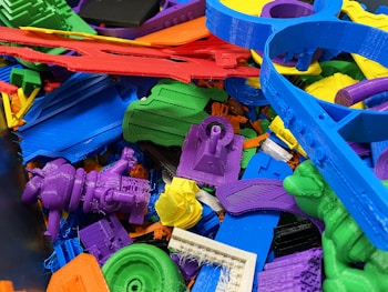 A collection of colorful 3D printed plastic objects in various shapes and colors, piled together. The objects include intricate designs such as gears, abstract forms, and tools, made from materials resembling different parts and components.