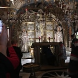 The image captures an ornate altar in an elaborate religious setting, featuring silver statues and a crucifix. Several individuals are engaged in prayer or taking photos, showing a mix of reverence and modern interaction with the sacred space. The interior is richly decorated with chandeliers and intricate artwork, creating an atmosphere of solemnity and devotion.