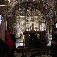 The image captures an ornate altar in an elaborate religious setting, featuring silver statues and a crucifix. Several individuals are engaged in prayer or taking photos, showing a mix of reverence and modern interaction with the sacred space. The interior is richly decorated with chandeliers and intricate artwork, creating an atmosphere of solemnity and devotion.
