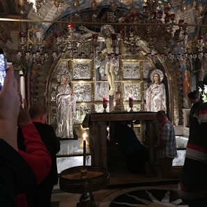 The image captures an ornate altar in an elaborate religious setting, featuring silver statues and a crucifix. Several individuals are engaged in prayer or taking photos, showing a mix of reverence and modern interaction with the sacred space. The interior is richly decorated with chandeliers and intricate artwork, creating an atmosphere of solemnity and devotion.