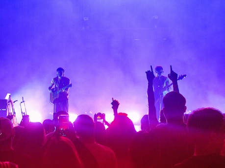 A concert scene features two performers on stage illuminated by purple and pink lighting. One is playing a guitar, while the other appears to be singing or speaking into a microphone. The silhouette of an enthusiastic audience is visible in the foreground, with some people raising their hands and capturing the moment on their phones.