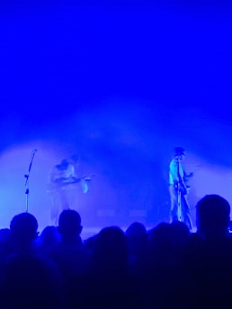 A concert scene with two musicians performing under a strong blue light. The musicians are playing guitars on stage with a crowd of silhouettes visible in front of them.