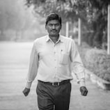 A man in business attire walks confidently down an outdoor pathway. The background is blurred, with trees and foliage lining the path. The image is in black and white, adding a timeless and serious feel.