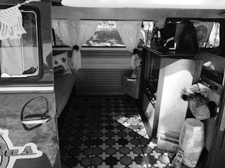The interior of a vintage van is styled with retro decor. Crocheted curtains hang over the windows, and a cushioned bench seat is adorned with decorative pillows. A vintage cabinet or wooden furniture piece is visible, along with baskets and a container holding artificial flowers. The patterned floor is reminiscent of mid-century design, adding to the nostalgic atmosphere.