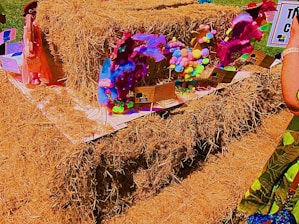 A collection of colorful, handcrafted sculptures made from various materials like pompoms and fabric is displayed on a large stack of hay. The sculptures are set on individual platforms with informational cards. Bright and vibrant hues dominate the scene.