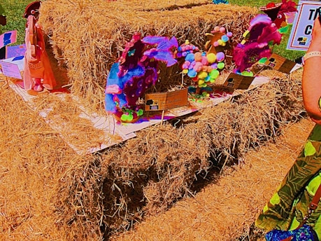A collection of colorful, handcrafted sculptures made from various materials like pompoms and fabric is displayed on a large stack of hay. The sculptures are set on individual platforms with informational cards. Bright and vibrant hues dominate the scene.