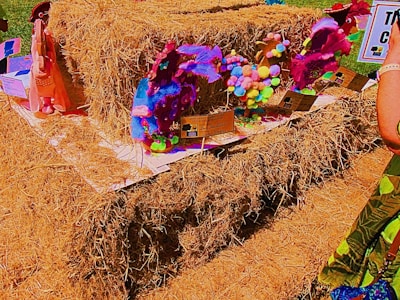 A collection of colorful, handcrafted sculptures made from various materials like pompoms and fabric is displayed on a large stack of hay. The sculptures are set on individual platforms with informational cards. Bright and vibrant hues dominate the scene.