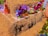 A collection of colorful, handcrafted sculptures made from various materials like pompoms and fabric is displayed on a large stack of hay. The sculptures are set on individual platforms with informational cards. Bright and vibrant hues dominate the scene.