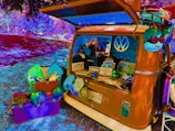 A vibrantly colored, psychedelic-style image features a vintage Volkswagen van parked in a park setting. The van's back is open, displaying various playful and quirky items such as plush toys, novelty signs, and decorative objects. An array of bright, colorful items are scattered around, including a purple box and vibrant masks or hats. The surrounding environment is surreal, with distorted color effects applied to the grass and trees.