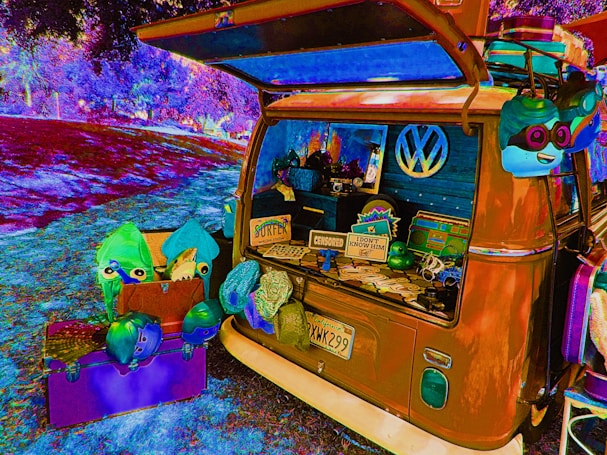 A vibrantly colored, psychedelic-style image features a vintage Volkswagen van parked in a park setting. The van's back is open, displaying various playful and quirky items such as plush toys, novelty signs, and decorative objects. An array of bright, colorful items are scattered around, including a purple box and vibrant masks or hats. The surrounding environment is surreal, with distorted color effects applied to the grass and trees.