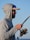 A person wearing a light gray hoodie and a cap is holding a fishing rod. The sun reflects on their sunglasses, and they appear to be focused on fishing. The background shows a clear blue sky.