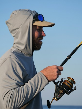 Bearded angler in a gray sun hoodie with the hood up under a cap, blue mirrored sunglasses pushed up on the brim, holding a spinning rod against a clear blue sky
