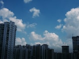 A cluster of high-rise buildings with modern architectural designs is set against a backdrop of a vivid blue sky populated with fluffy white clouds. A construction crane is visible on one building, indicating ongoing development.