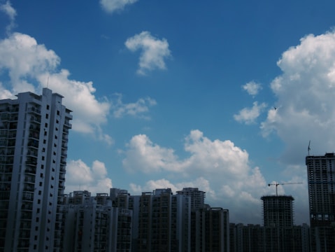 A cluster of high-rise buildings with modern architectural designs is set against a backdrop of a vivid blue sky populated with fluffy white clouds. A construction crane is visible on one building, indicating ongoing development.
