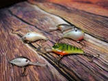 Five colorful fishing lures with multiple hooks are arranged on a wood-textured surface, displaying intricate details and varied color patterns.