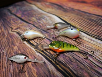 Five colorful fishing lures with multiple hooks are arranged on a wood-textured surface, displaying intricate details and varied color patterns.