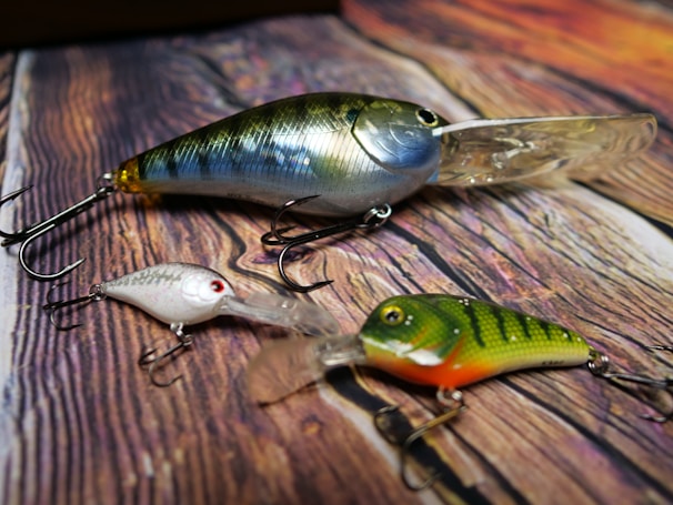 Three fishing lures of varying sizes and colors are arranged on a textured wooden surface. The lures have shiny, reflective finishes and realistic features including scales, eyes, and hooks.