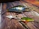 Three fishing lures of varying sizes and colors are arranged on a textured wooden surface. The lures have shiny, reflective finishes and realistic features including scales, eyes, and hooks.
