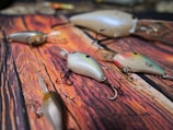 A collection of artificial fishing lures with multiple hooks is spread out on a wooden surface. The lures vary in color, featuring shades of pearl, pink, green, and silver, with realistic detailing resembling small fish. Each lure is equipped with sharp treble hooks, demonstrating their design for catching fish.
