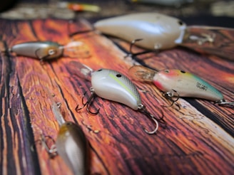 A collection of artificial fishing lures with multiple hooks is spread out on a wooden surface. The lures vary in color, featuring shades of pearl, pink, green, and silver, with realistic detailing resembling small fish. Each lure is equipped with sharp treble hooks, demonstrating their design for catching fish.