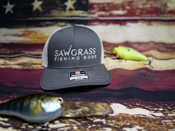 A gray and white baseball cap with the words 'Sawgrass Fishing Rods' on the front is placed on a rustic, weathered American flag backdrop. In front of the cap are two fishing lures, one green and yellow, the other blue and silver.