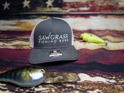 A gray and white baseball cap with the words 'Sawgrass Fishing Rods' on the front is placed on a rustic, weathered American flag backdrop. In front of the cap are two fishing lures, one green and yellow, the other blue and silver.