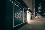 A city street at night with a modern glass building facade showcasing a large management company logo. The area is illuminated by streetlights, and a parking garage entrance is visible on the right. The street is quiet, with some lights visible in the windows of a large building in the background.